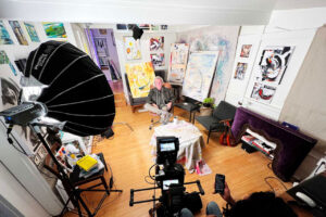 Behind the scenes, a camera setup filming an interview of Ben young sitting in his studio surrounded by his paintings