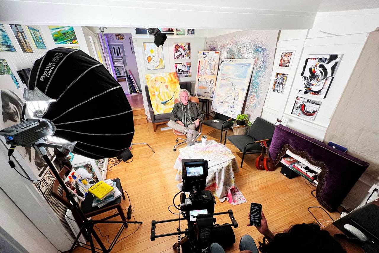 Behind the scenes, a camera setup filming an interview of Ben young sitting in his studio surrounded by his paintings