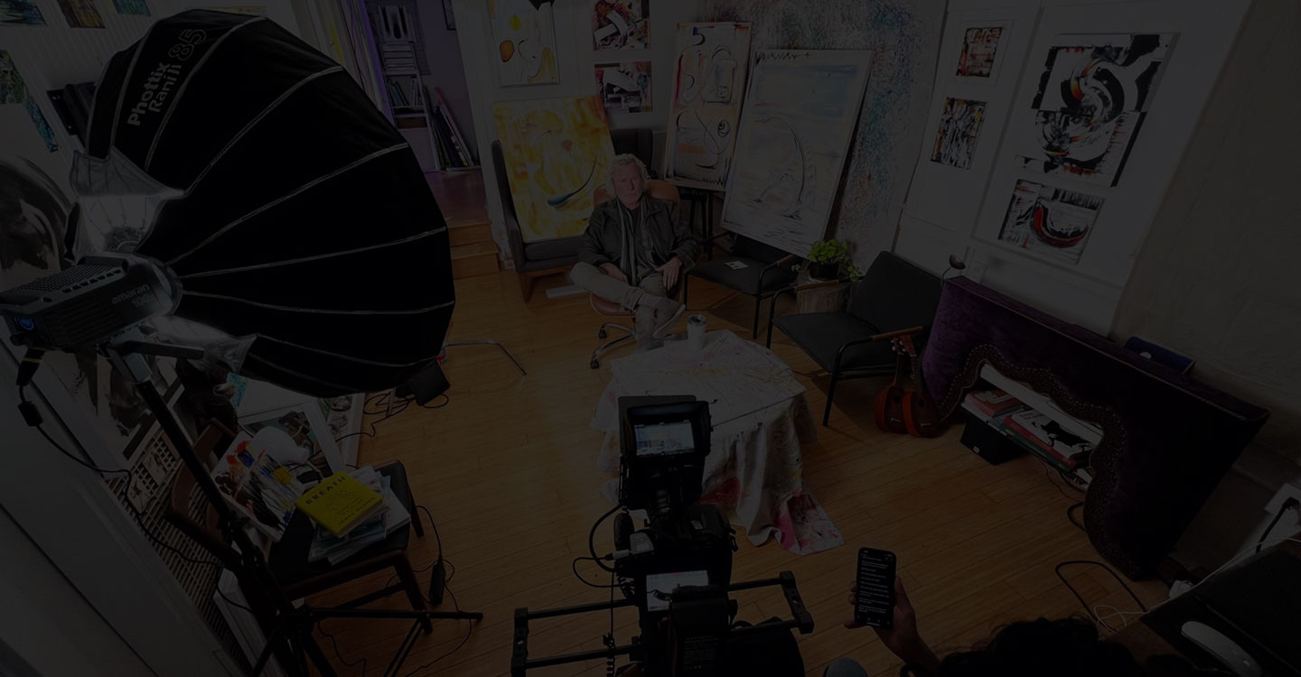 Behind the scenes, a camera setup filming an interview of Ben young sitting in his studio surrounded by his paintings