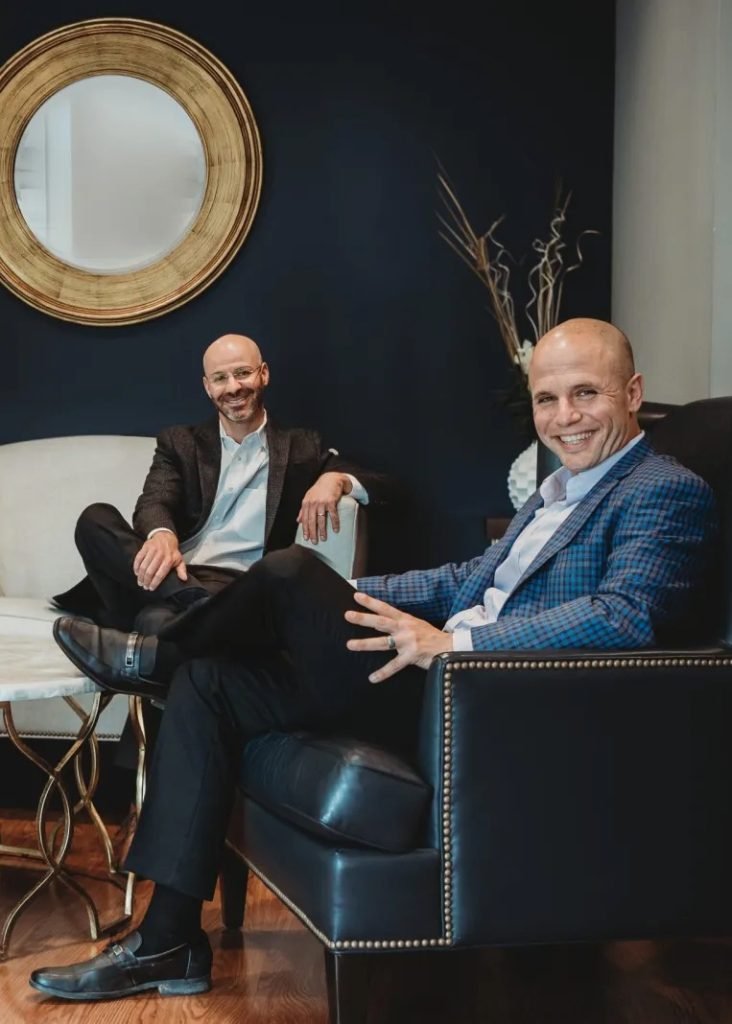 Two men in business attire sit in a modern office lounge—one in a blue blazer on a black leather chair in the foreground, the other in a dark blazer on a white sofa behind him—both smiling, with a round wall mirror and decorative vase on a dark accent wall. MindShift Financial Coaching case study corporate brand film by 20EVO