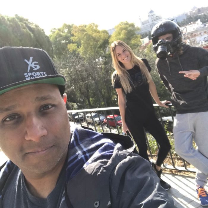 man taking a selfie with actors in the background one of them wearing a motorcycle helmet with a camera attached