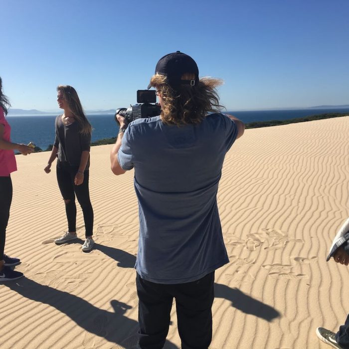 Man directing shoot on beach with multiple models with ocean in the background.