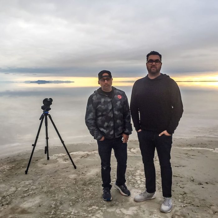 two men standing facing the camera while sun is setting with another camera pointing at the sunset with lake in the background